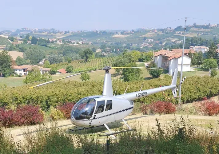 A silver Robinson R44 helicopter with registration I-GYWD parked in a scenic vineyard landscape.
