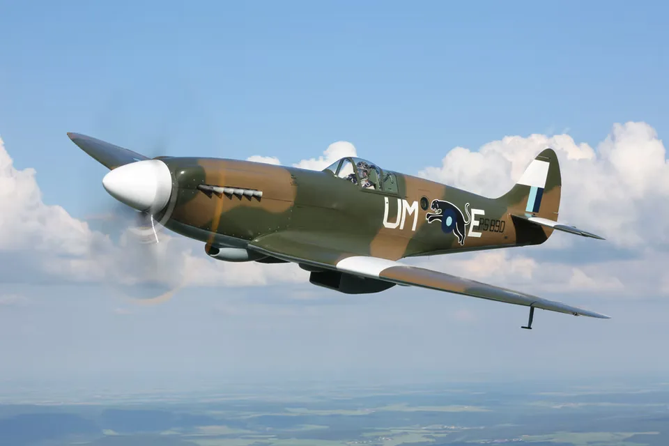 A Supermarine Spitfire in a camouflage paint scheme with 'UM-E S690' markings flying against a blue sky with clouds.