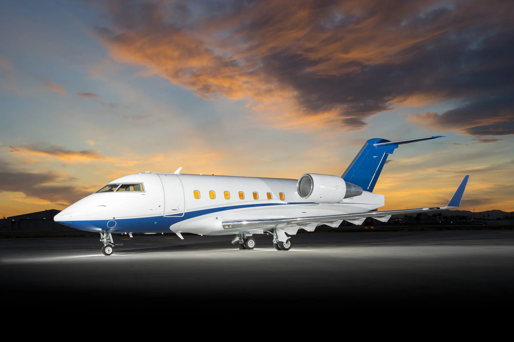A white business jet with blue accents on the nose and tail, parked on a tarmac at sunset.