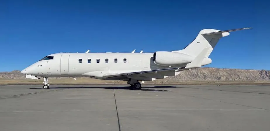 A white business jet is parked on an airfield with mountains in the background under a clear blue sky.