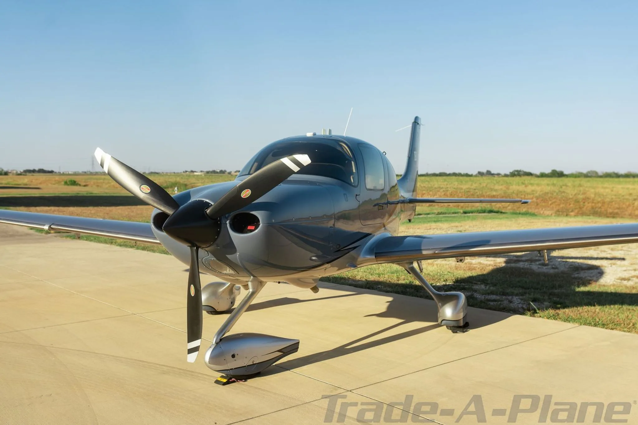 A front-quarter view of a grey Cirrus SR22 aircraft with a three-blade propeller on a tarmac.