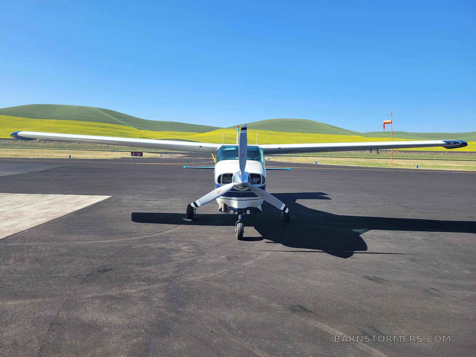 A front view of a white and blue Cessna T206H Turbo Stationair parked on an airfield with yellow fields and green hills in the background.