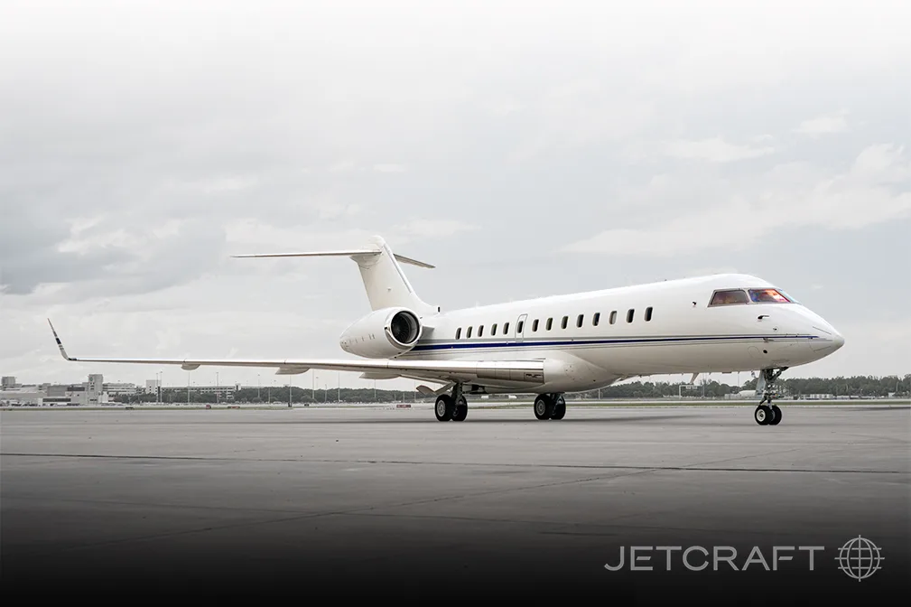 A white Bombardier Global Express business jet with blue and grey stripes on the tarmac under a cloudy sky.