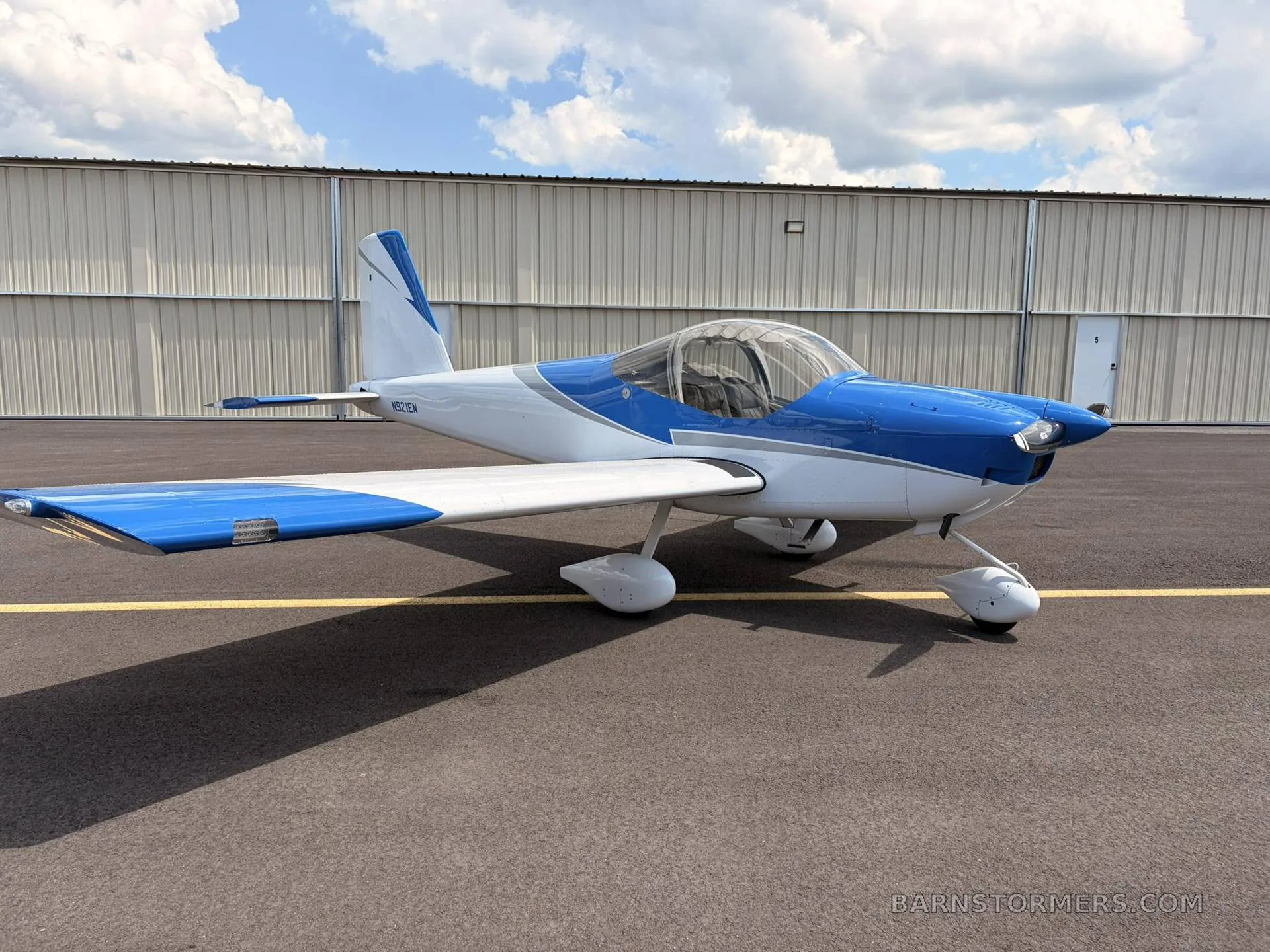A blue and white single-engine aircraft with registration N921EN, parked on an asphalt tarmac in front of a hangar.
