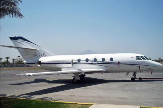 A white Dassault Falcon 50 business jet with blue stripes is parked on an tarmac under a clear sky, with mountains in the background.