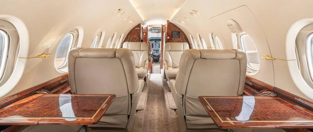 A wide view of the business jet's elegant cabin interior, featuring beige leather seating, polished wood tables, and a spacious aisle.
