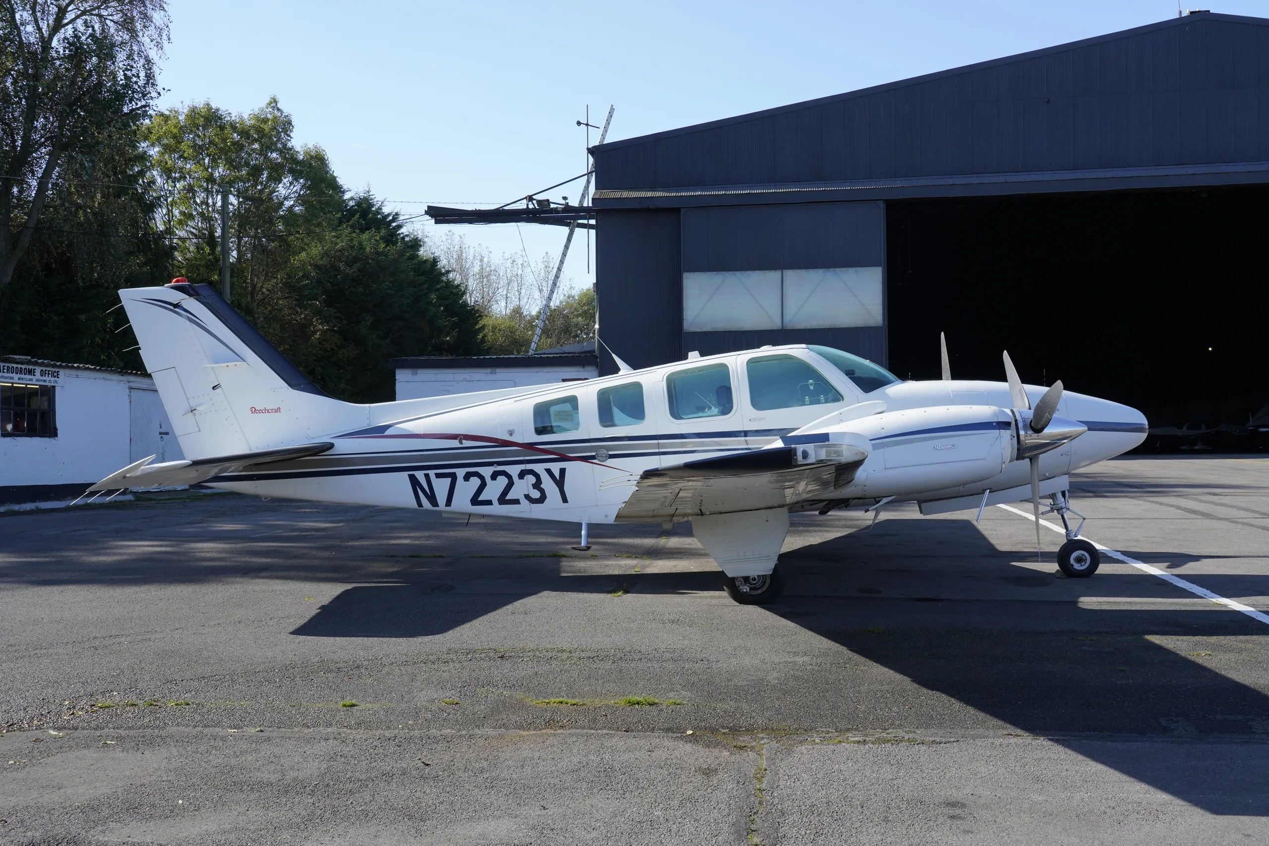 A white twin-engine propeller aircraft with blue and red stripes, registration N7223Y, parked on an asphalt apron next to a hangar.