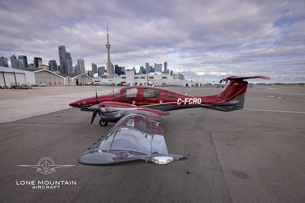 A red and grey Diamond DA62 aircraft with registration C-FCRQ parked on an airfield with the Toronto skyline in the background.