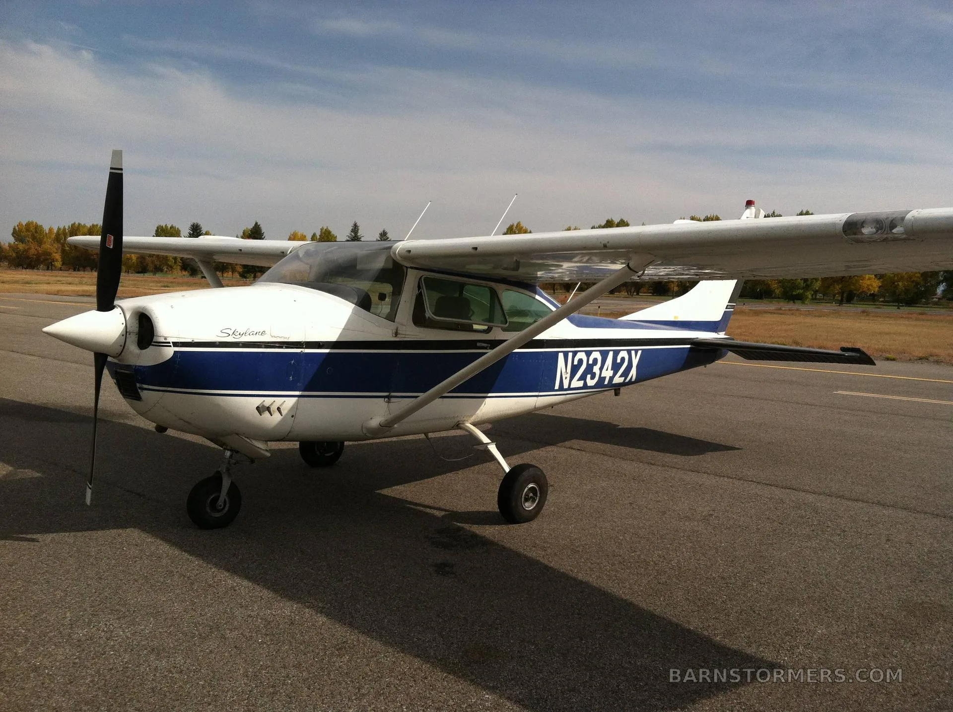 A white and blue Cessna 182 Skylane (N2342X) parked on an asphalt runway with autumn trees in the background.