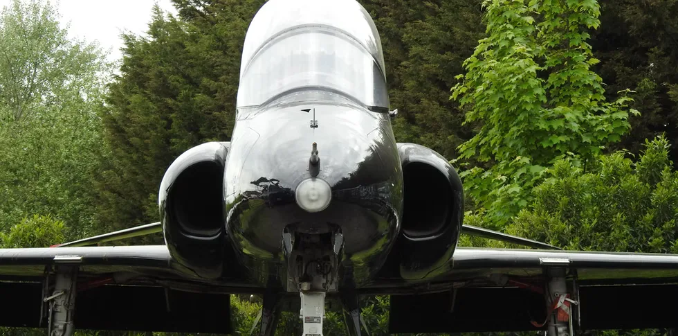 Front view of a black BAE Hawk jet aircraft parked on the ground, surrounded by green trees.