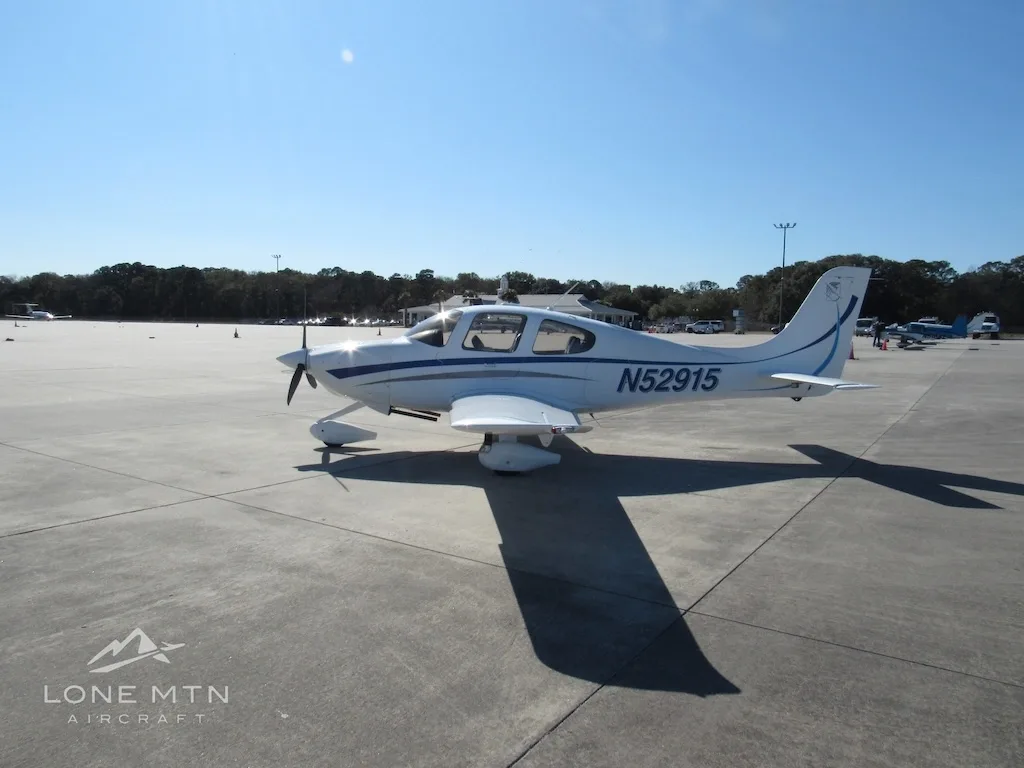 A white Cirrus SR20 aircraft with blue and grey stripes is parked on a concrete ramp, viewed from the front-right.