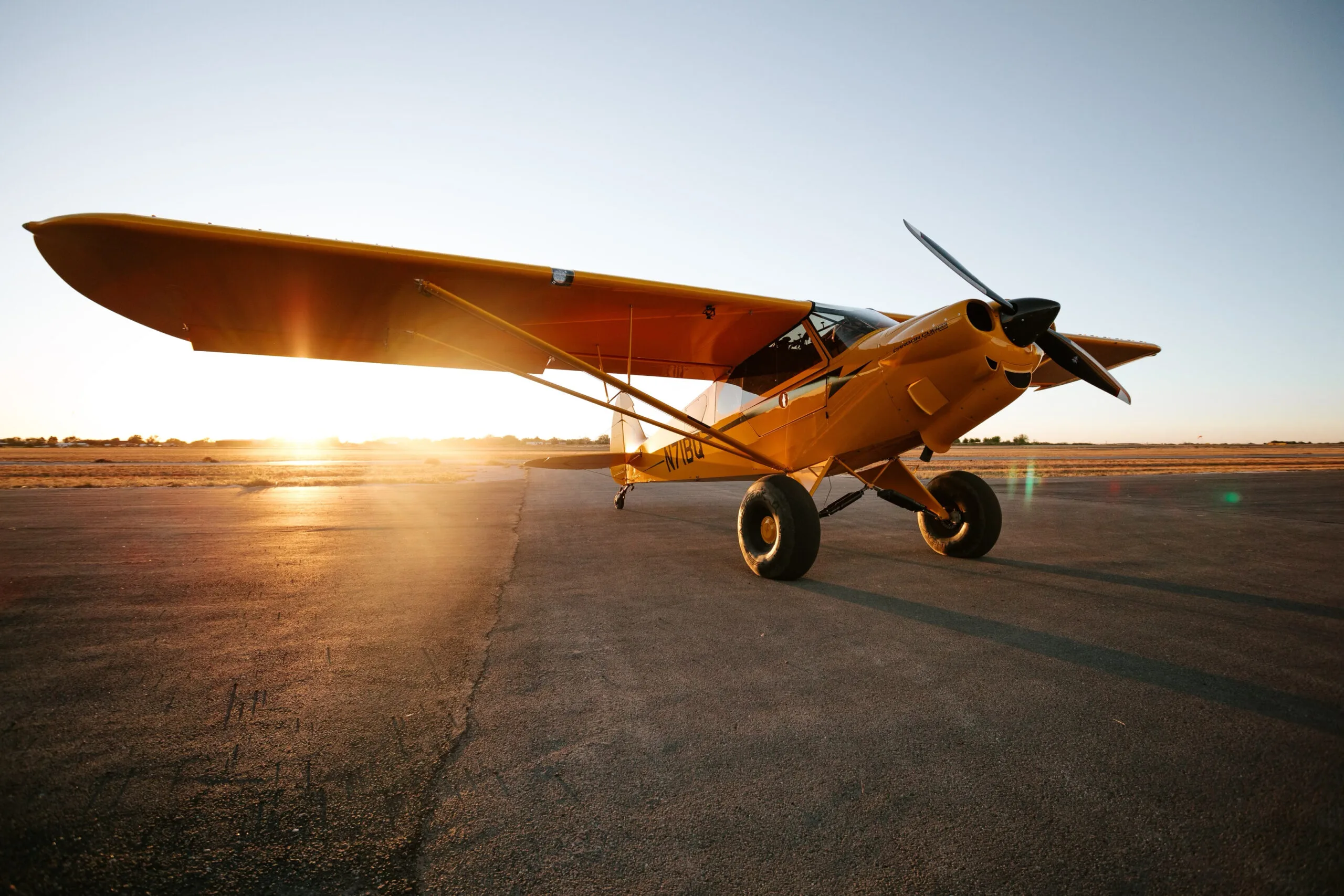 A yellow Carbon Cub aircraft with large tires is parked on an airfield at sunset, with the sun low on the horizon.