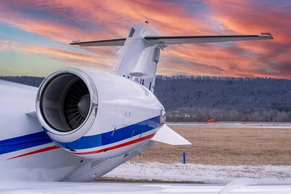 A rear-quarter view of the tail and engine of a white Learjet 45 with blue and red stripes, under a vibrant sunset sky.