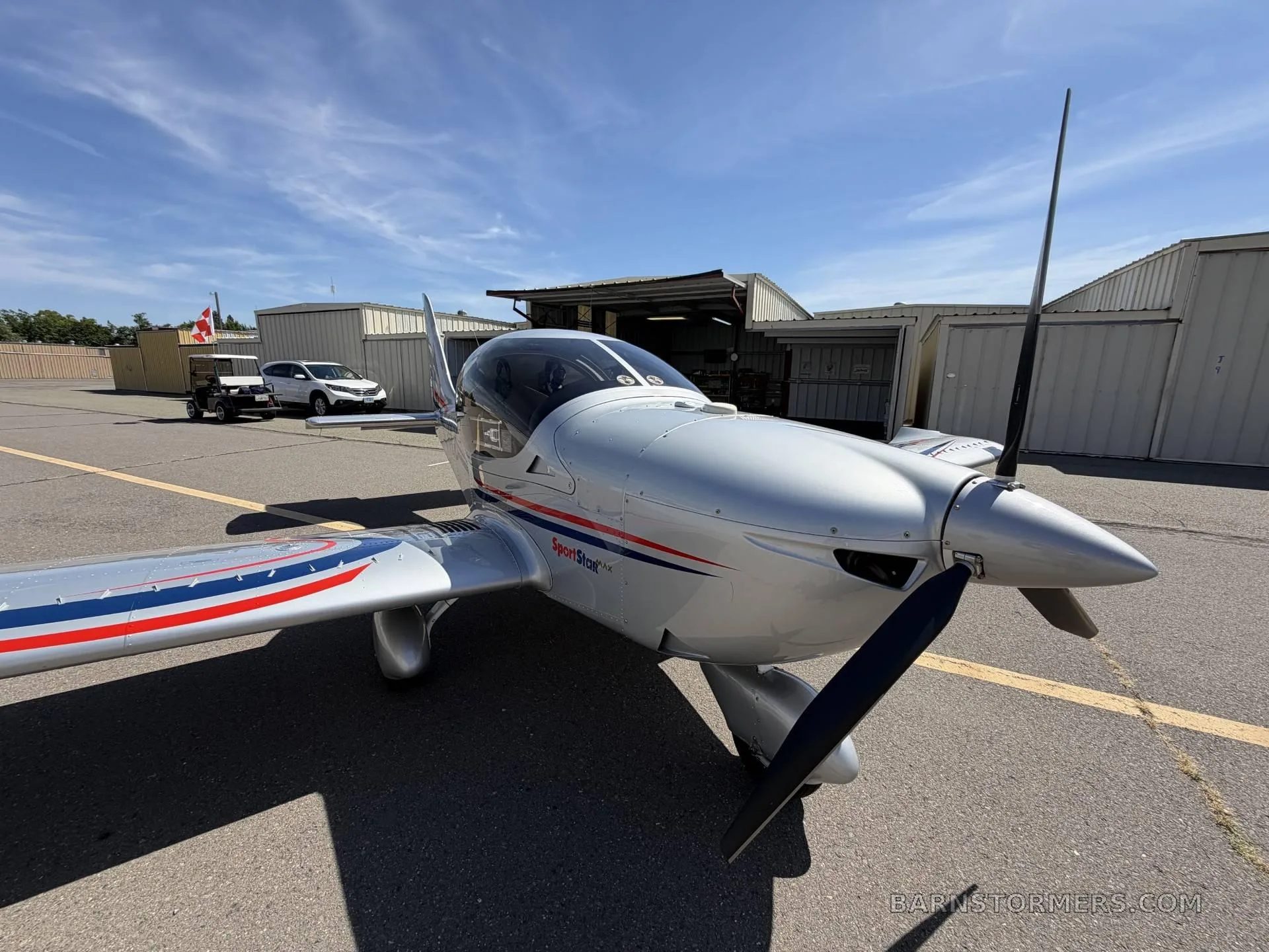 A front-right view of the silver SportStar MAX Turbo aircraft with red and blue stripes, parked on an asphalt tarmac in front of hangars.
