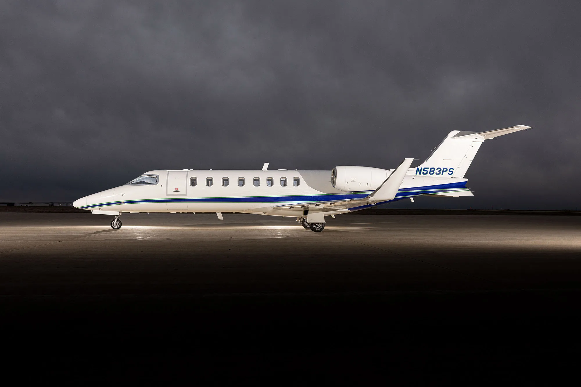 A white Learjet 75 with blue and green stripes, registration N583PS, parked on an tarmac under dramatic cloudy skies.