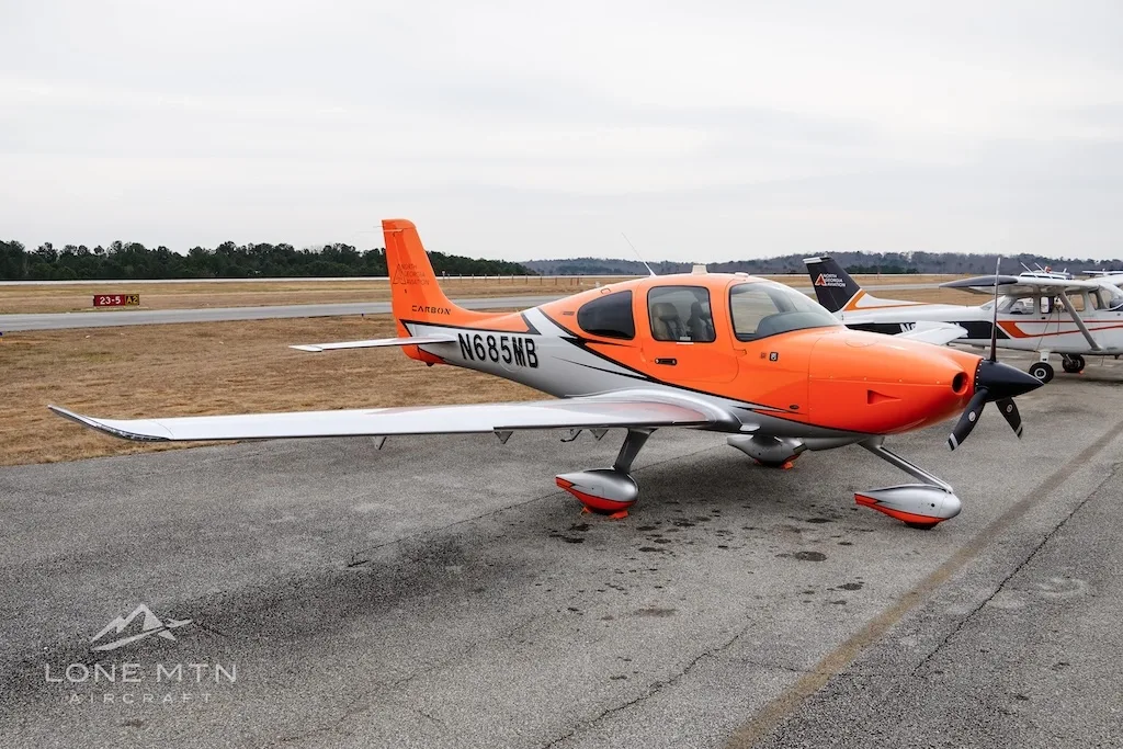 The orange and silver Cirrus SR20 aircraft N685MB parked on an airfield with other planes in the distance.