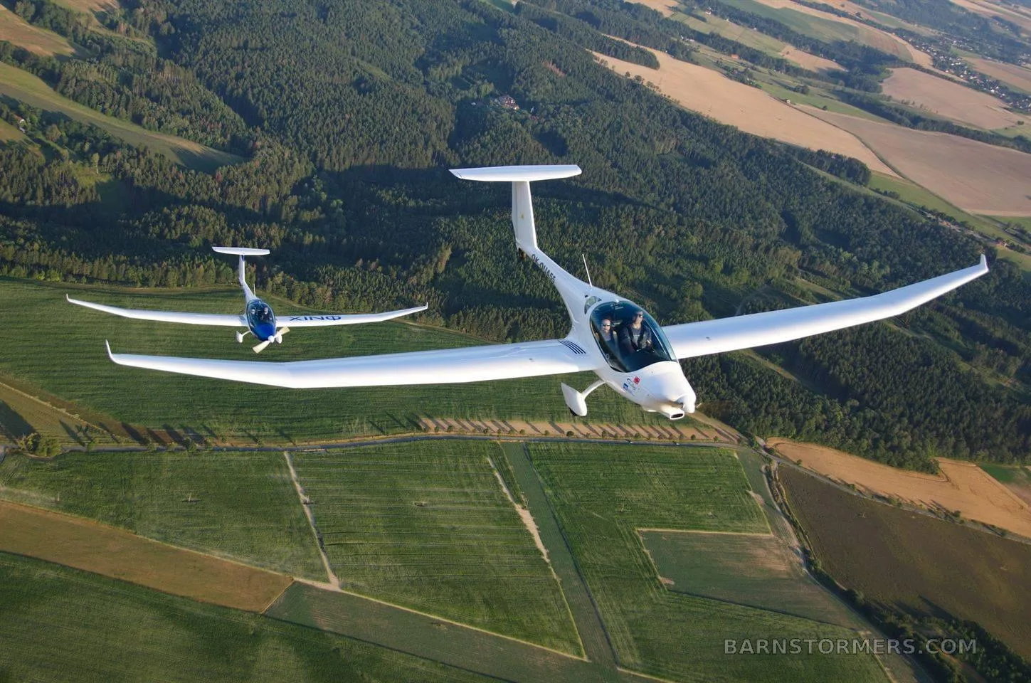 Two white motor gliders fly in formation over a lush green and forested landscape.