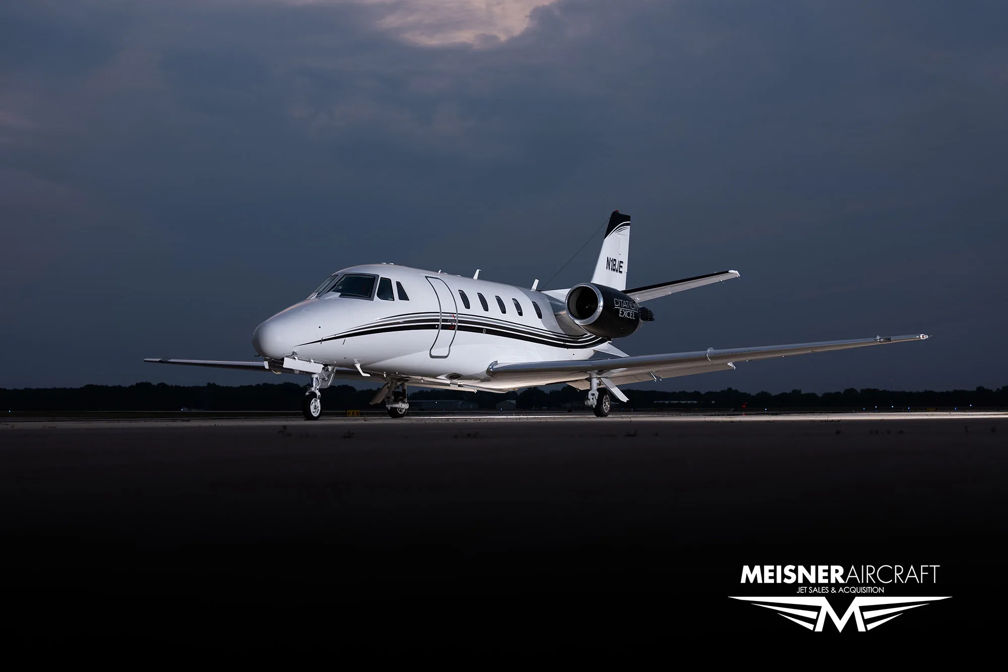 A white Citation Excel business jet with black and white stripes on the tail and engines, parked on a tarmac at dusk, viewed from the front-left.
