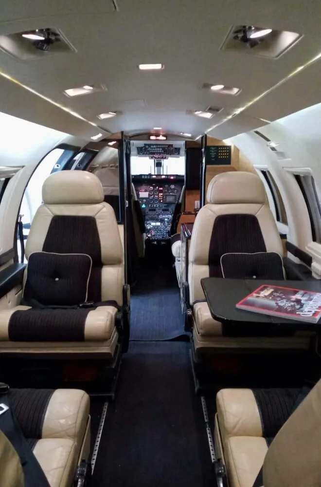 Interior view of a private jet cabin looking forward towards the cockpit, with beige and dark brown seats and a table.