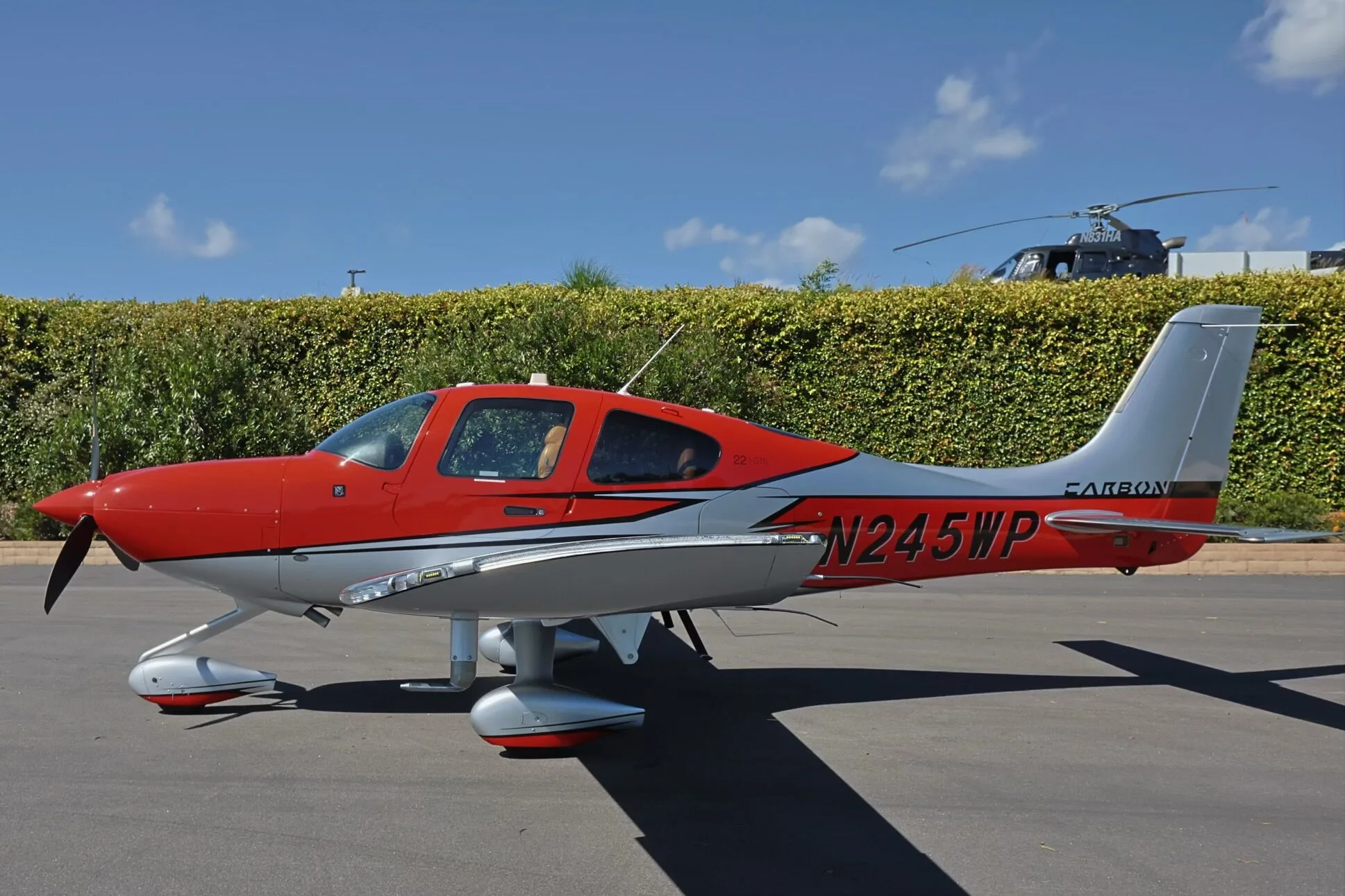A red and silver Cirrus SR22 GTS aircraft, N245WP, parked on an tarmac with a helicopter in the background.