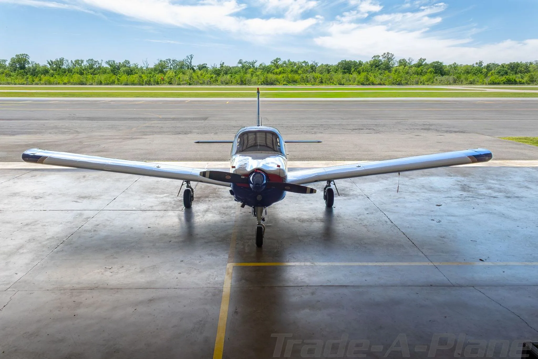 Front view of a blue and white Piper Arrow II aircraft positioned at a hangar entrance, facing a runway.