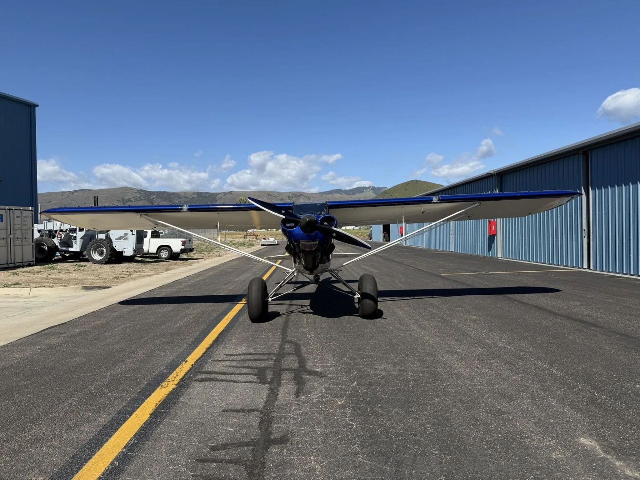 A blue and white Carbon Cub SS aircraft with large tundra tires, seen head-on on an airport runway with hangars in the background.