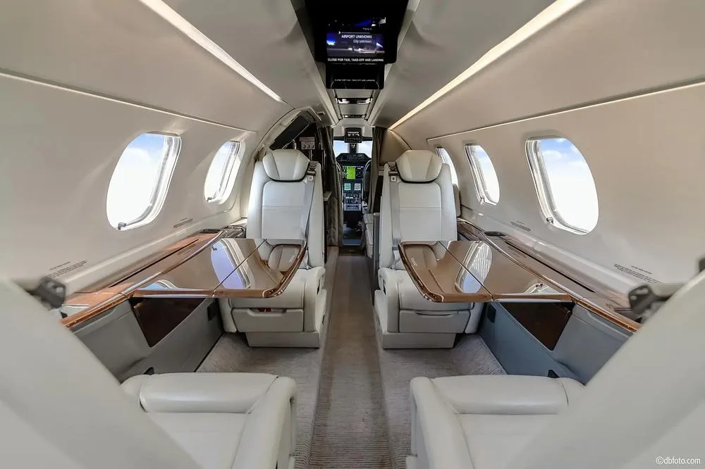 The luxurious interior cabin of a private jet, featuring light-colored seats, wooden tables, and overhead screens.