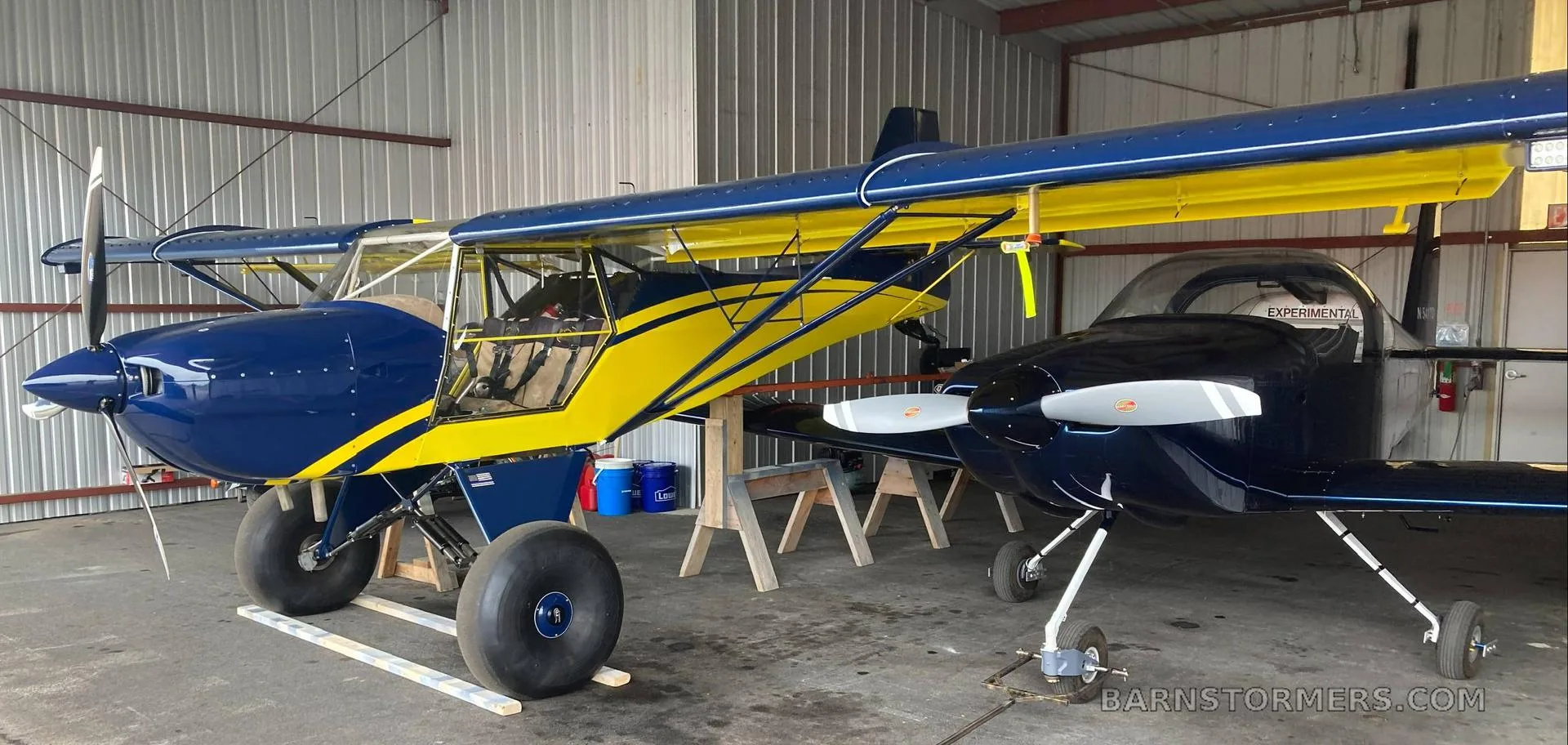 A blue and yellow high-wing aircraft with tundra tires parked in a hangar next to another dark blue aircraft.