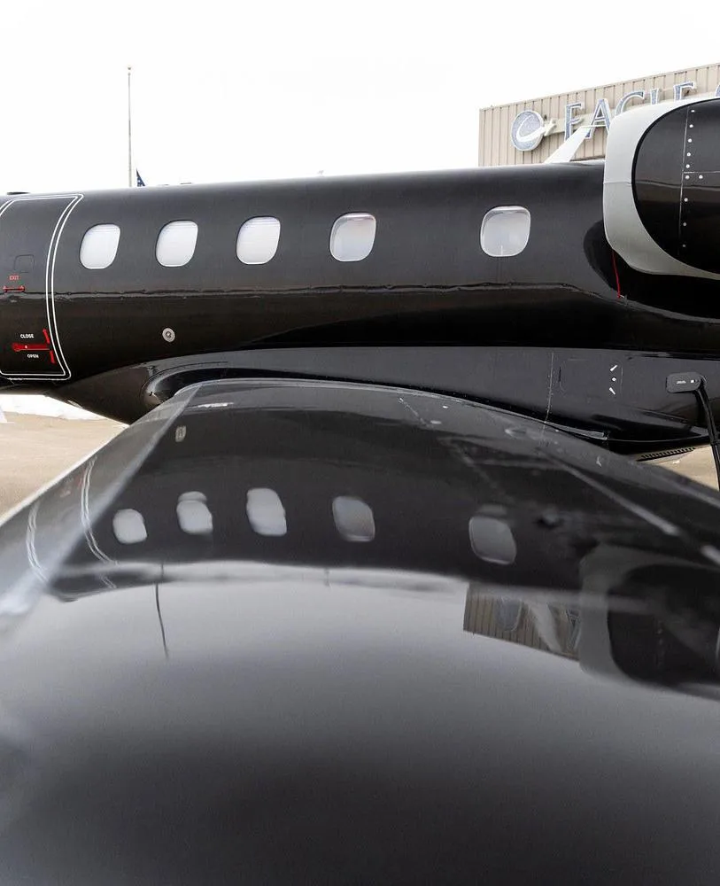 A close-up view of the mid-fuselage and wing of a black Embraer Phenom 300 jet, showing passenger windows and reflections.