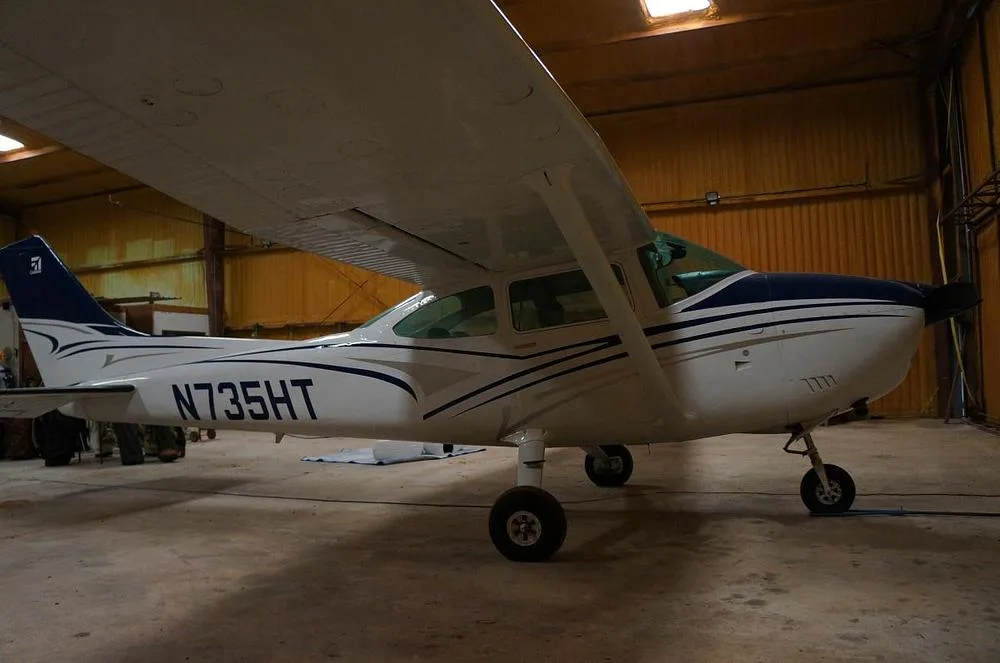 A white and blue Cessna 182 Skylane N735HT is parked in a hangar, viewed from the right side.