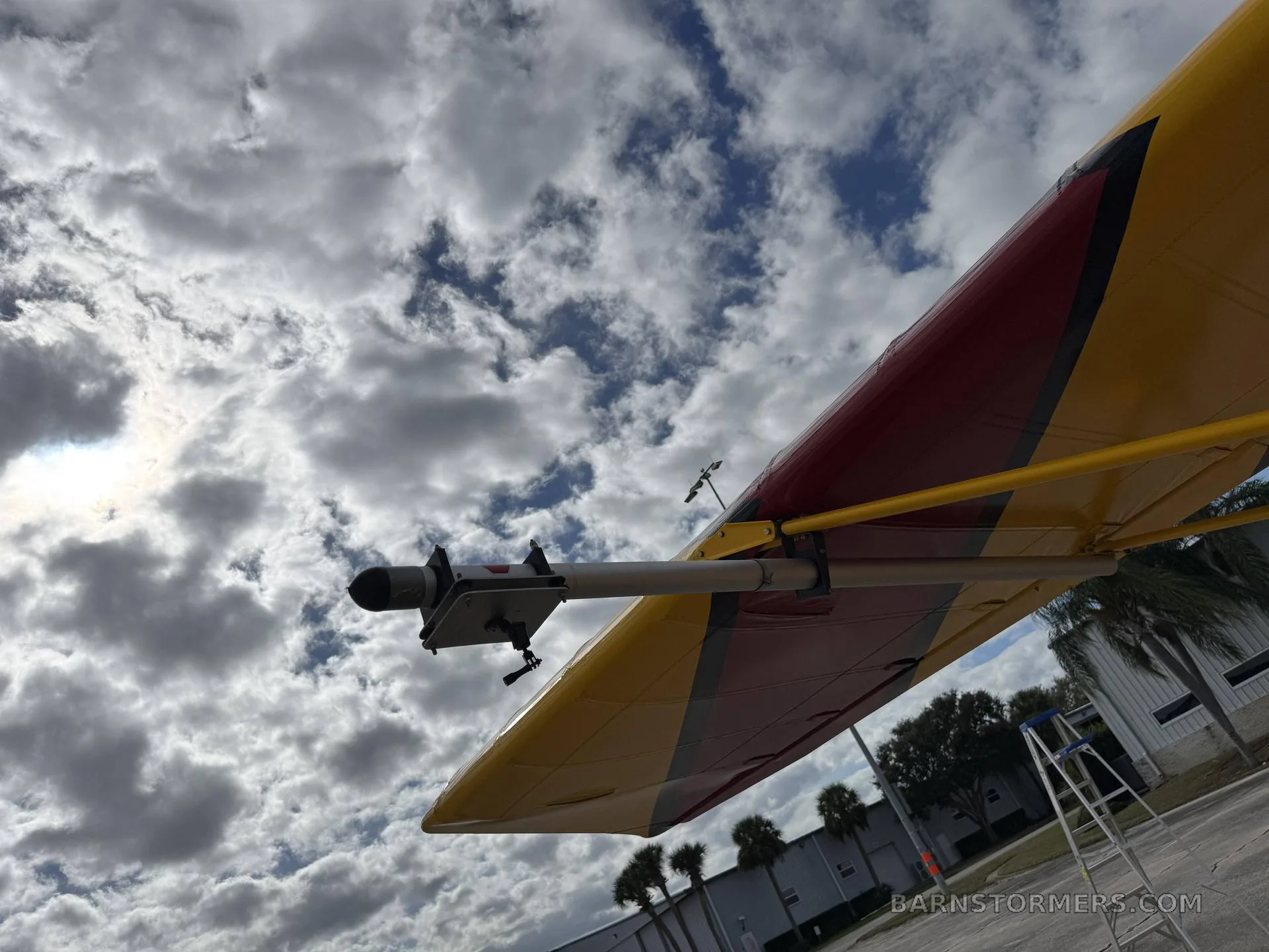 A low-angle view looking up at the yellow and red wing of an Air-Cam seaplane, showing a mounted sensor or camera pod against a cloudy sky.