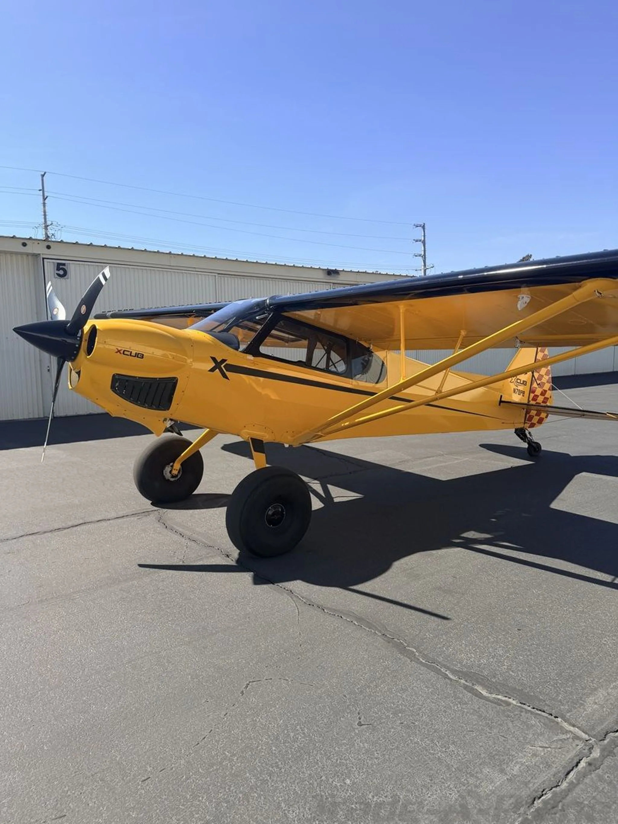 A yellow CubCrafters XCub with black wings and large tires is parked on an asphalt tarmac next to a hangar.