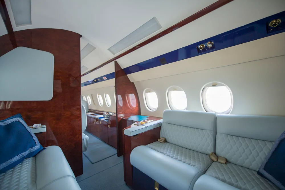 The rear section of the private jet's cabin, featuring a light grey sofa with blue pillows, dark wood paneling, and multiple windows.