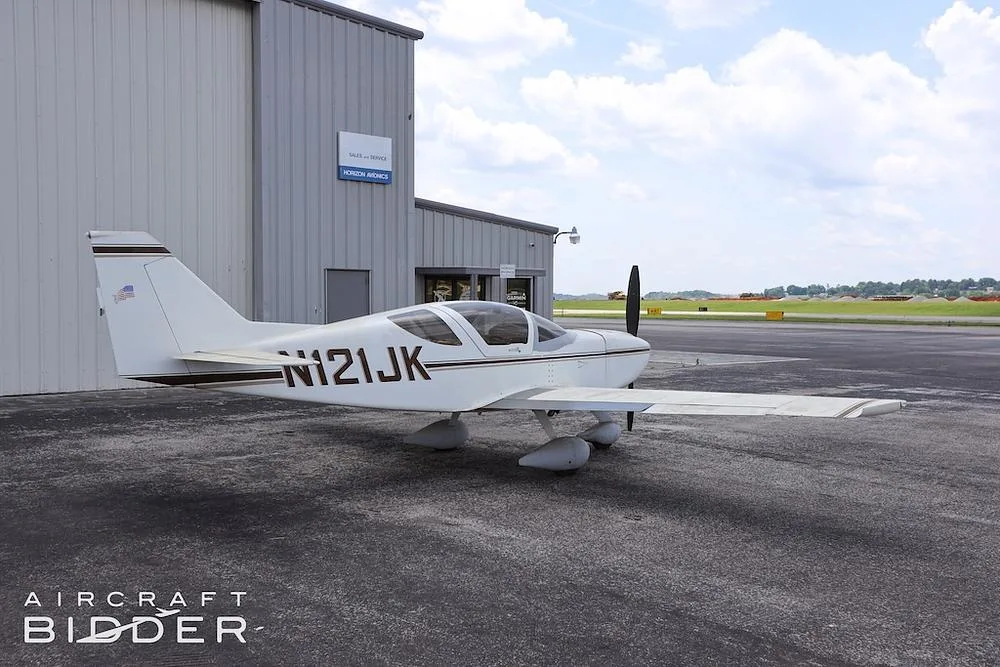 A white single-engine aircraft (N121JK) with brown stripes parked on an asphalt apron next to a hangar.