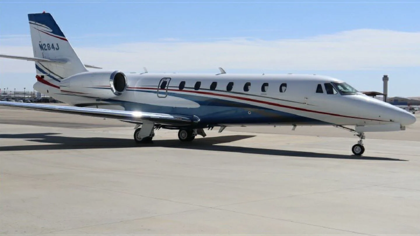 A white business jet with blue and red stripes, tail number N284J, parked on an airport tarmac under a clear sky.
