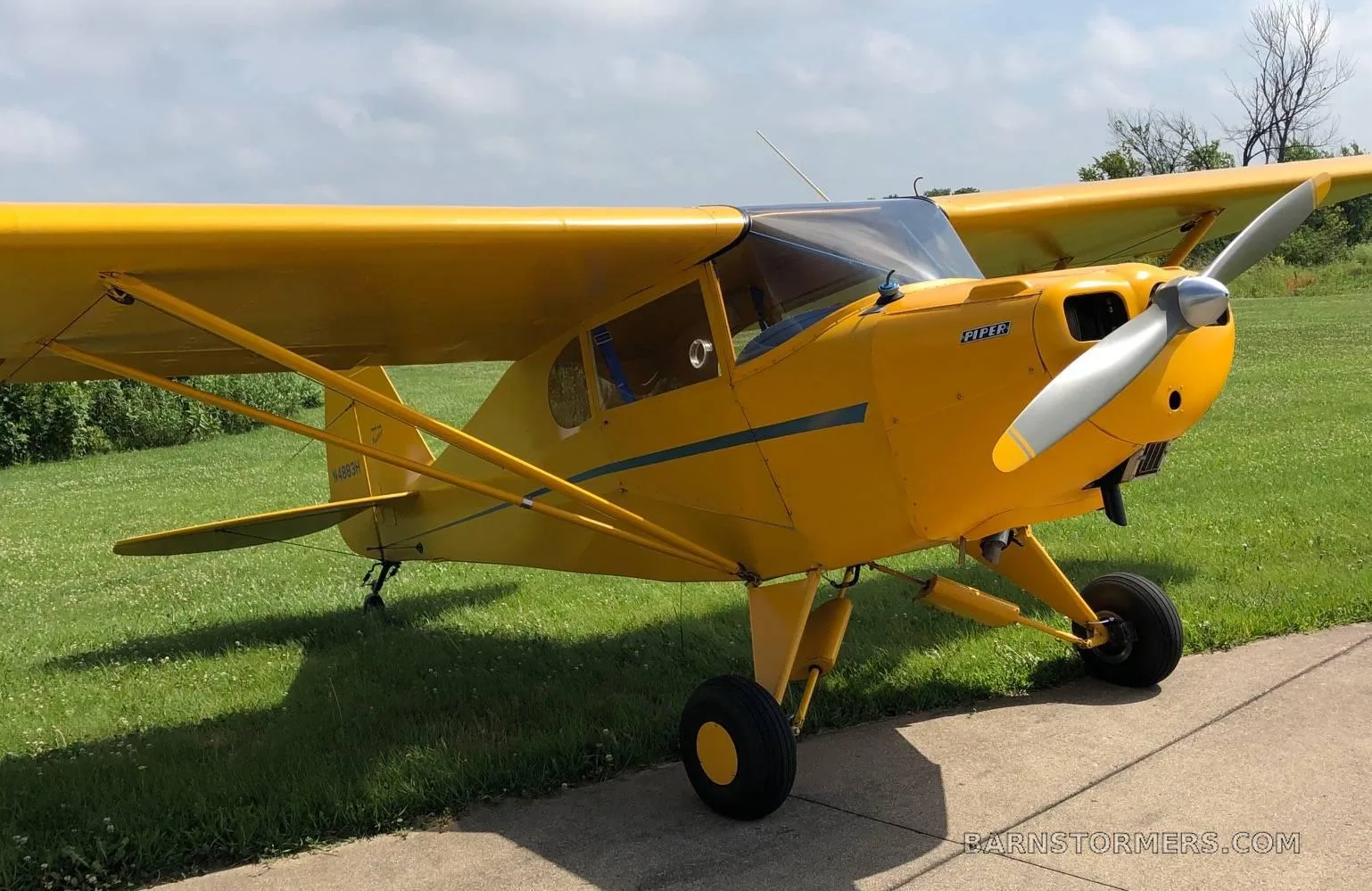 A yellow Piper Cub aircraft with a blue stripe, parked on grass and concrete, seen from the front-right.