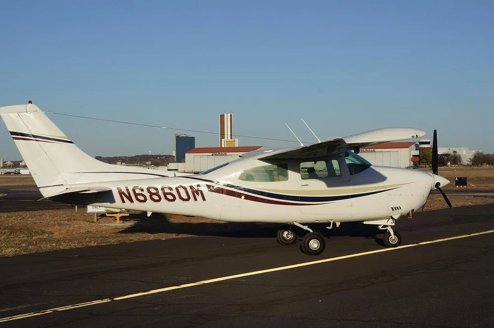 A white Cessna 210 Centurion aircraft with red, blue, and gold stripes parked on an airfield with buildings and a city skyline in the distance.