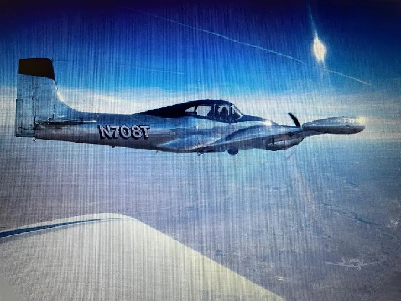 A polished silver single-engine aircraft with tail number N708T flies in a clear blue sky, viewed from another aircraft's wing.