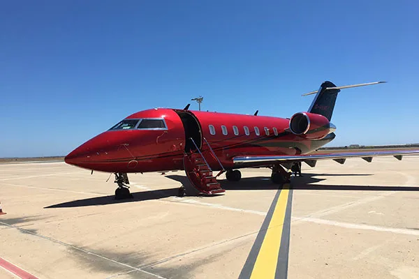 A metallic red Bombardier Challenger business jet with its main door open and stairs extended, parked on an airport tarmac under a clear blue sky.