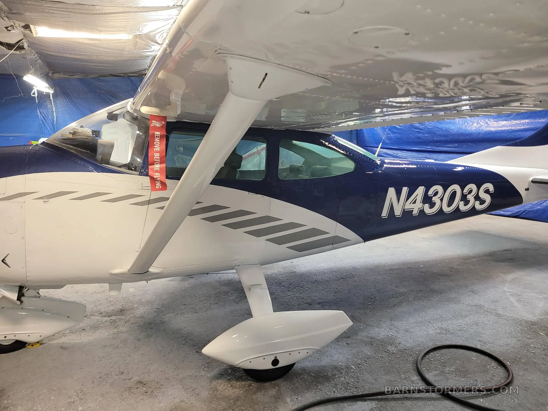 Side view of a blue and white aircraft with tail number N4303S, showing a 'REMOVE BEFORE FLIGHT' tag on the wing strut.