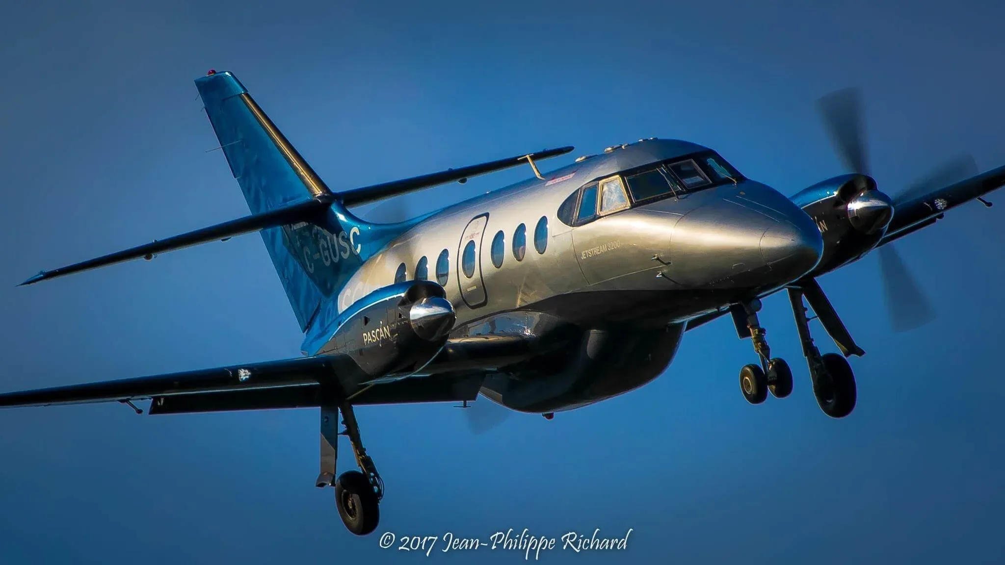 A silver and blue British Aerospace Jetstream 3200 turboprop aircraft, registered C-GUSC, in flight against a clear blue sky.
