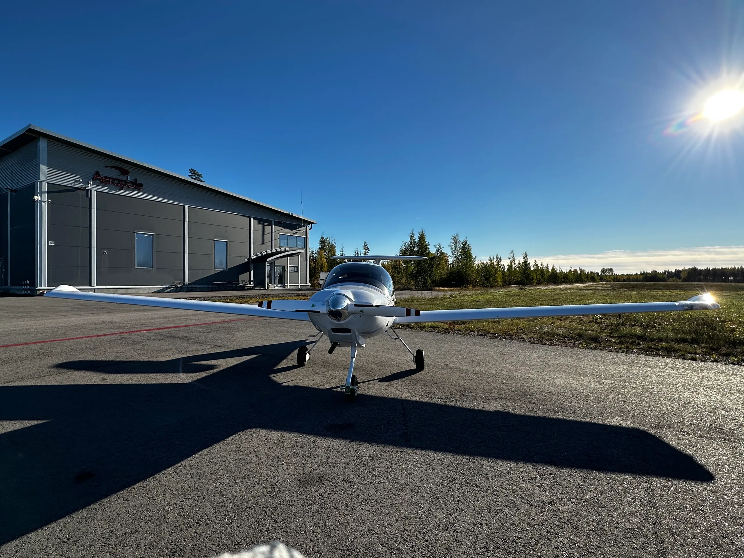 A white Diamond DA20 aircraft with red and black stripes parked on an asphalt apron, facing a hangar under a bright sun.