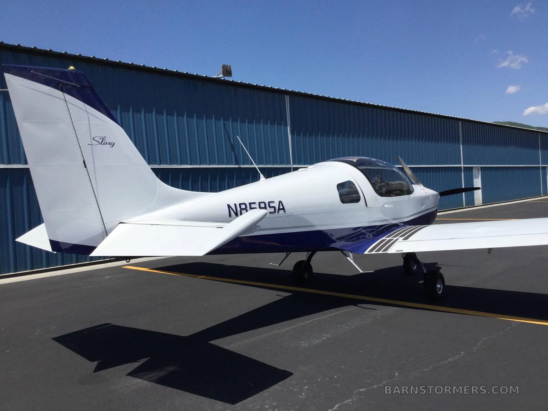 A white and blue Sling aircraft with registration N859SA is parked on an asphalt next to a blue hangar, viewed from the rear-left.