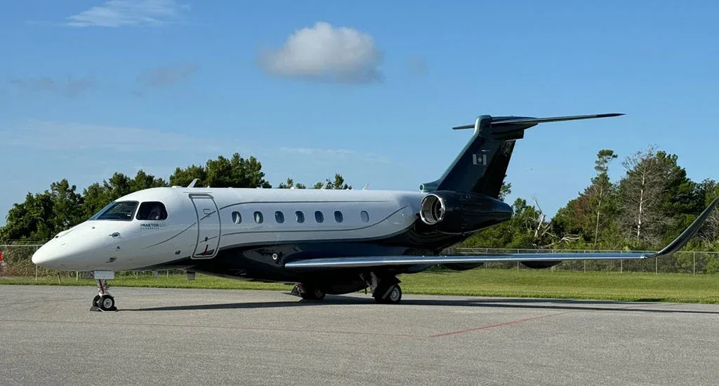 A white and dark grey Embraer Praetor 600 business jet with a Canadian flag on the tail, parked on an airfield under a blue sky.