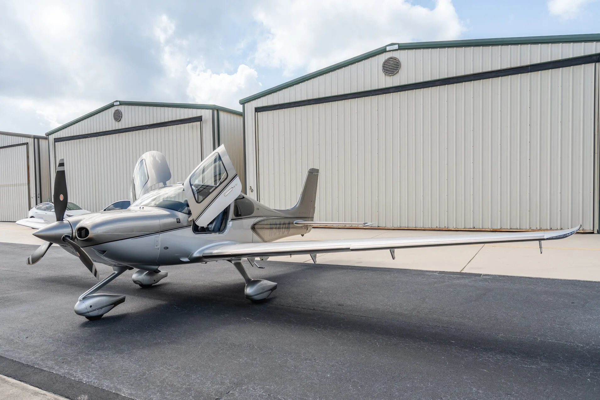 Three-quarter front view of a silver Cirrus SR22T aircraft (N3404) with its gull-wing doors open, parked on an airport tarmac.