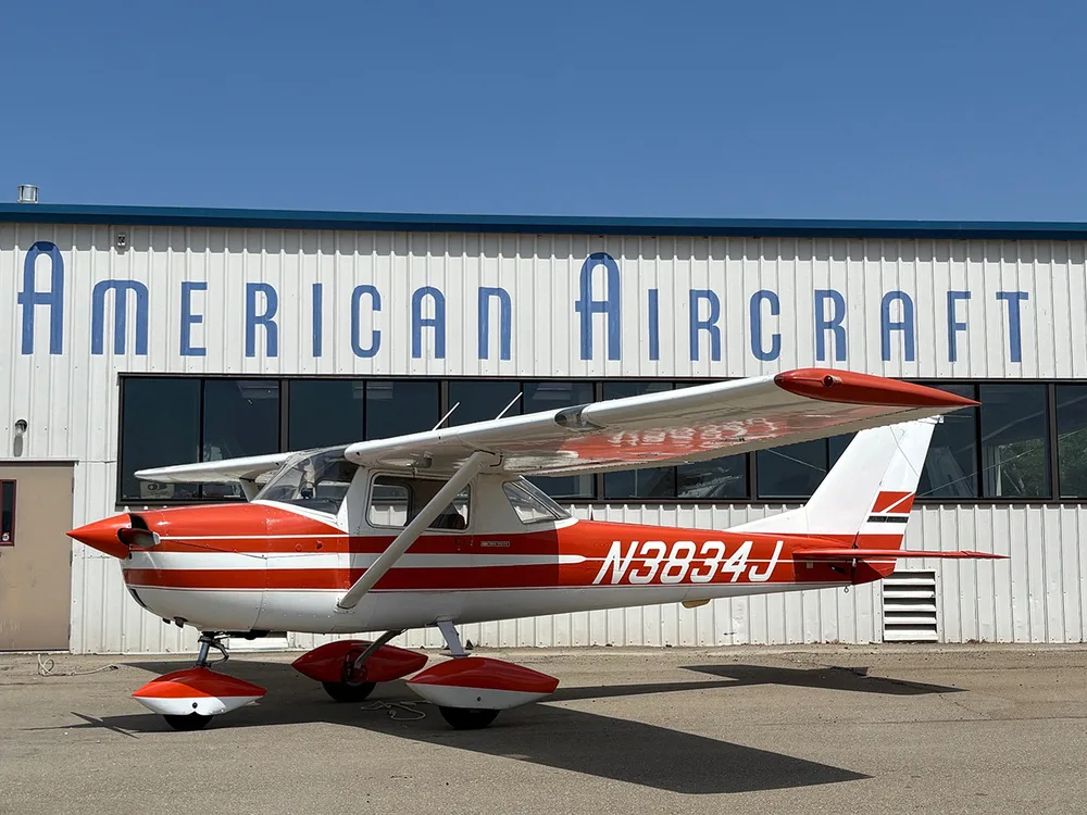 A red and white Cessna 172 aircraft with registration N3834J parked on an airfield in front of an "American Aircraft" hangar.