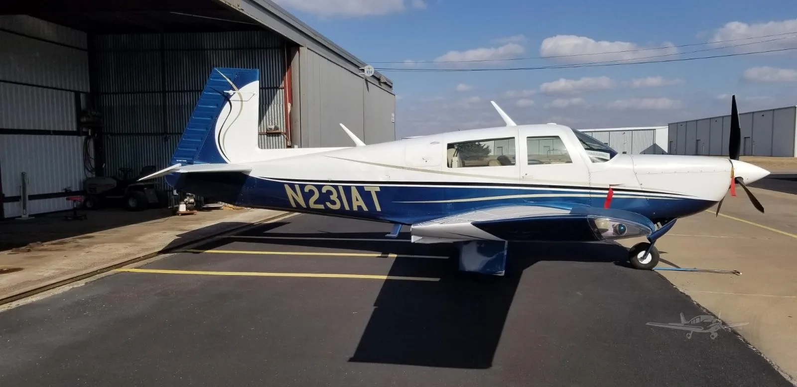 A white and blue single-engine propeller aircraft with gold accents is parked on an asphalt ramp outside a hangar.