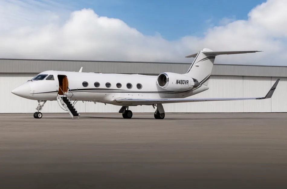 A white Gulfstream business jet with black stripes is parked on an tarmac with its main door open and stairs extended, in front of a hangar.