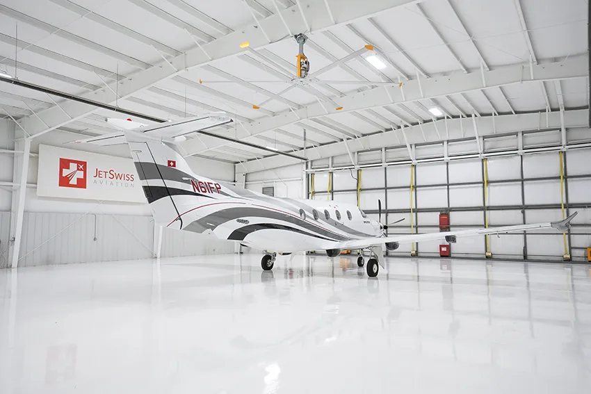 A white Pilatus PC-12 aircraft with gray and red stripes is seen from the rear in a brightly lit hangar.