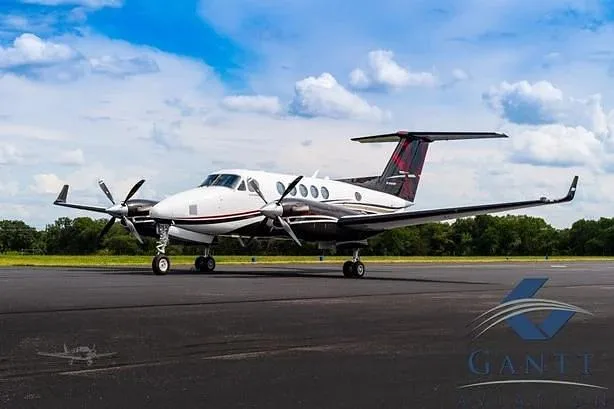 A white and dark brown twin-turboprop aircraft, likely a King Air, is parked on an asphalt runway under a partly cloudy sky.